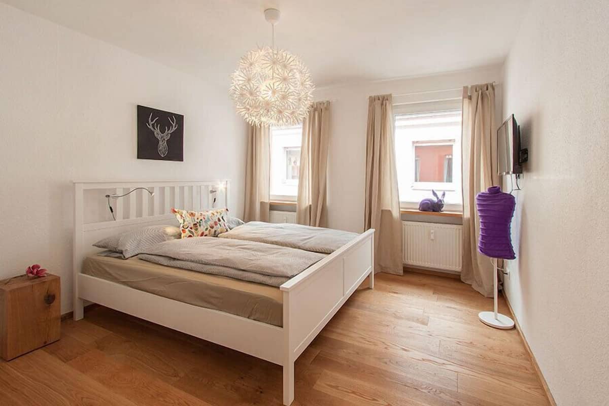 Une chambre à coucher avec un lit blanc, un luminaire décoratif au plafond, un téléviseur fixé au mur et une fenêtre avec des rideaux.