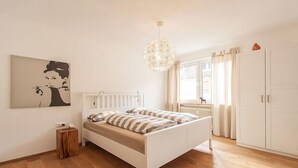 Une chambre à coucher comprenant un lit, un radiateur, un luminaire suspendu, une fenêtre avec des rideaux et une armoire.