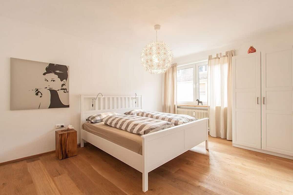 Une chambre à coucher comprenant un lit, un radiateur, un luminaire suspendu, une fenêtre avec des rideaux et une armoire.