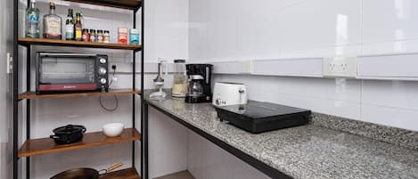 A kitchen with a microwave, coffee maker, toaster, and a shelf with bottles and a pot.