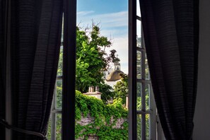 Une fenêtre ouverte avec des rideaux, une vue sur un bâtiment surmonté d’un dôme, et de la verdure.