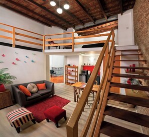 A loft-style living space with a wooden staircase, a dining area, and a kitchen visible through an open door.