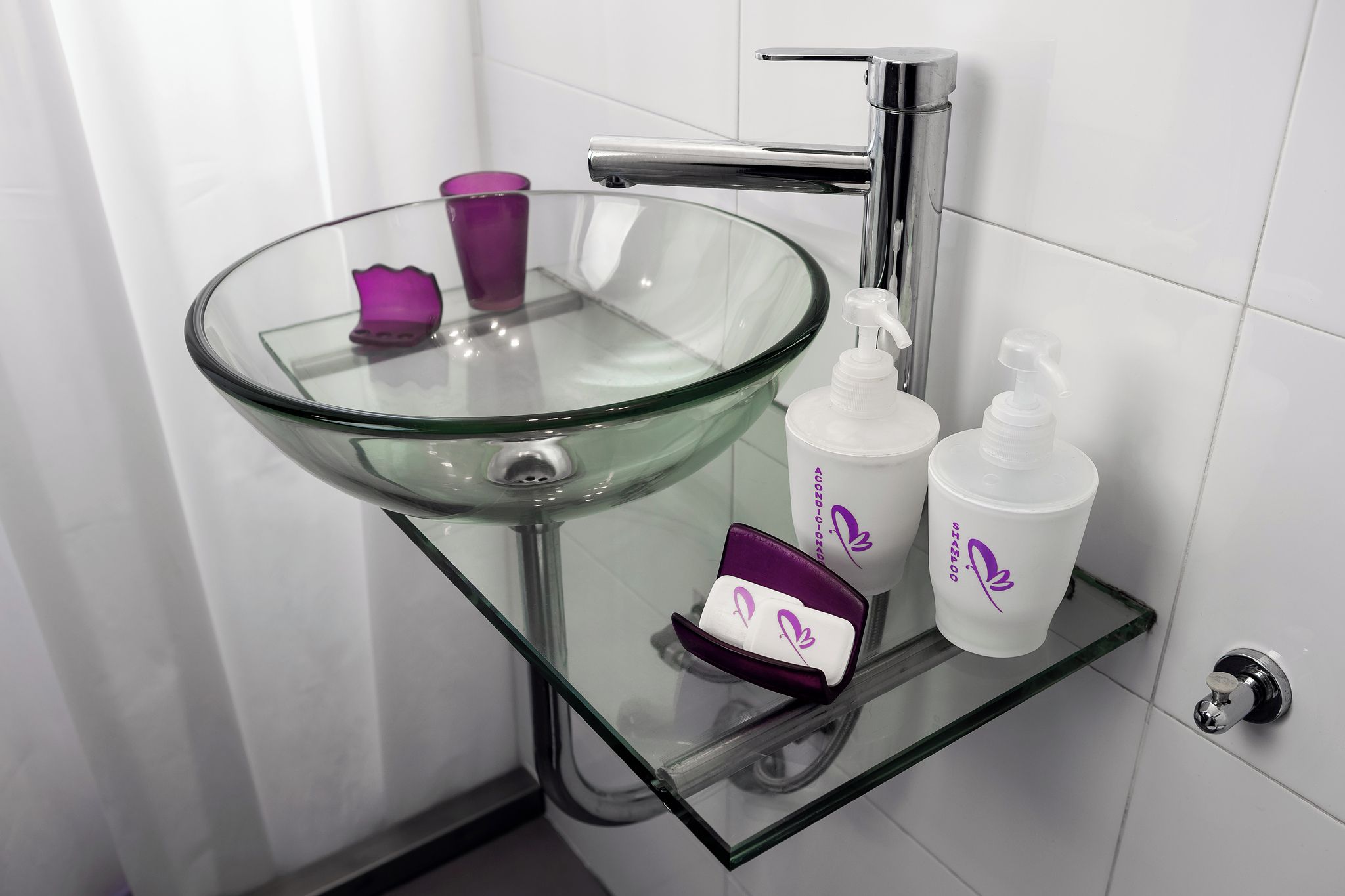A bathroom with a glass sink, a chrome faucet, and a glass shelf holding toiletries.