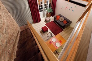 Un escalier mène à un espace de vie comprenant un canapé, un tapis rouge et une table à manger recouverte d’une nappe à pois.