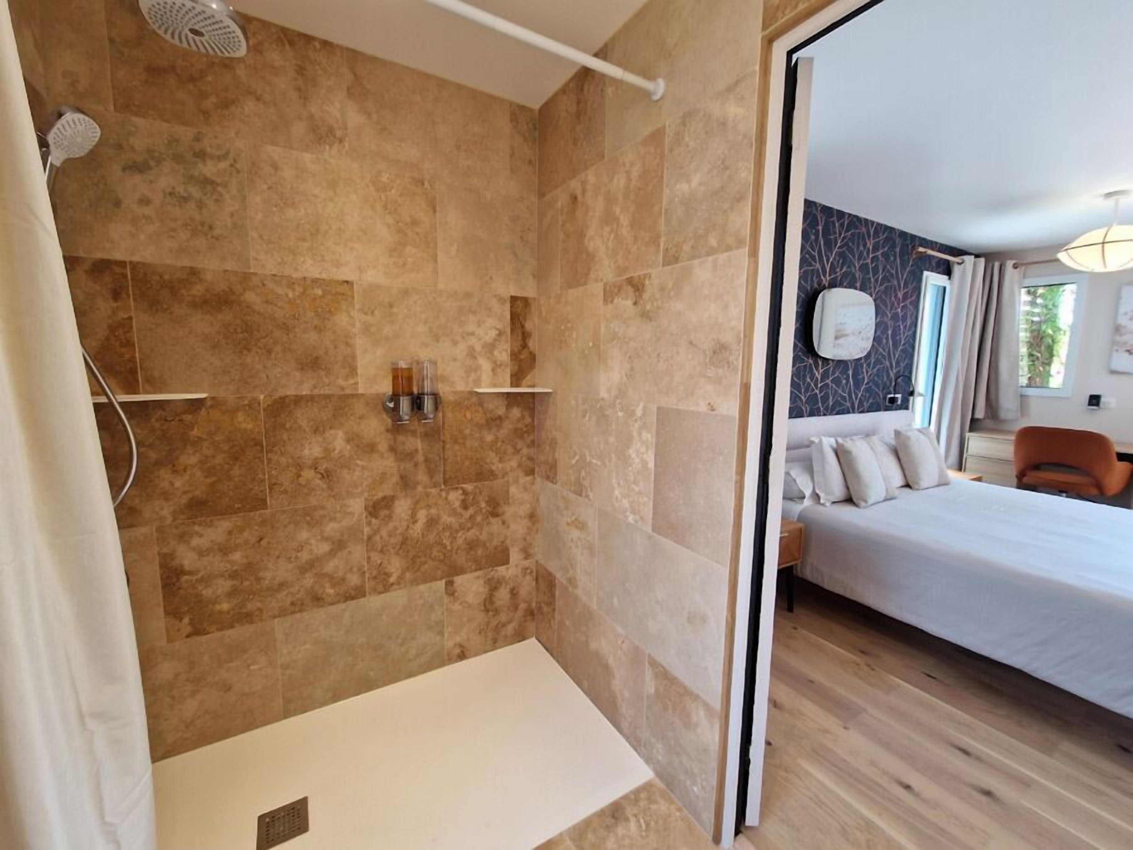 A bathroom with a shower, a toilet, and a sink. There is a bed with pillows and a nightstand in the adjacent room.