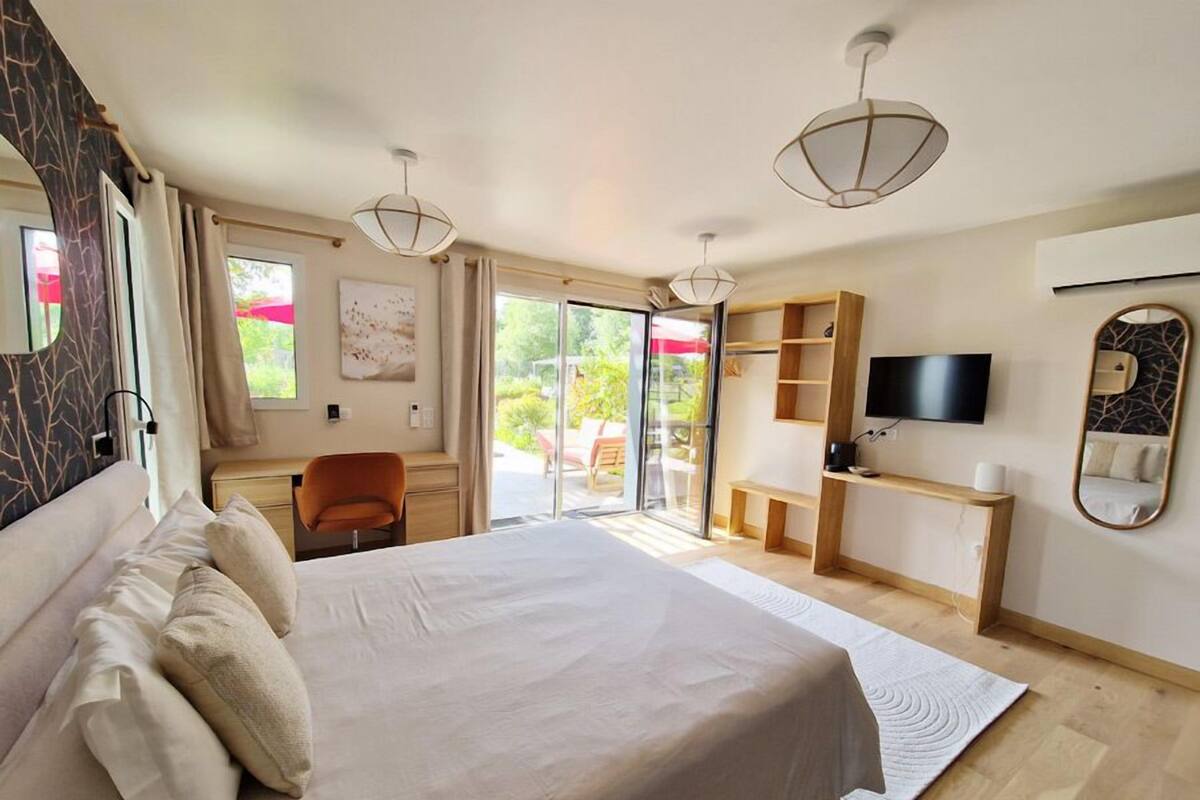 A bedroom with a large bed, a desk, a chair, a TV, and a sliding glass door to a balcony.