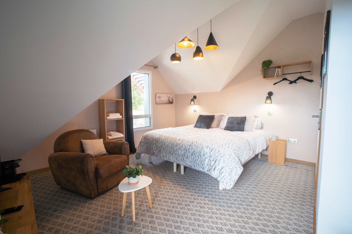 A bedroom with a sloped ceiling, a bed with a patterned bedspread, a brown armchair, a small table with a plant, and a window with a view.