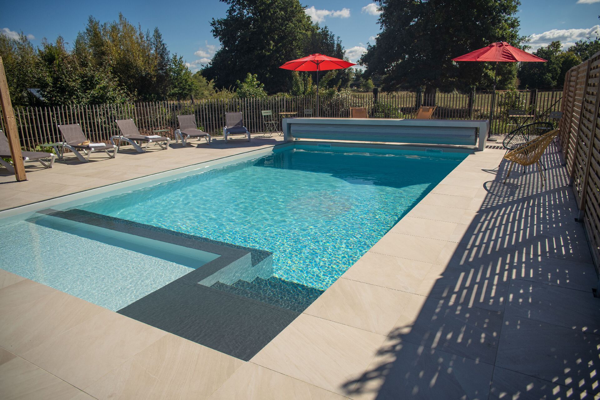 Une piscine rectangulaire à l’eau d’un bleu limpide, entourée d’une clôture en bois et ombragée par des parasols rouges.
