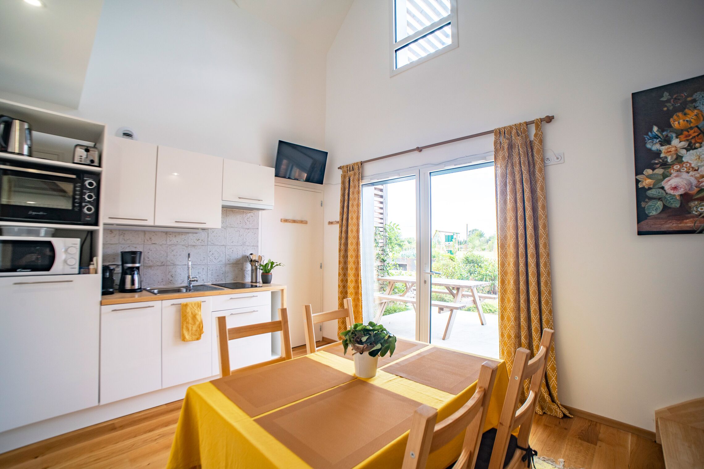A compact kitchen with white cabinets, a microwave, and a coffee maker. There is a dining table with chairs and a potted plant.