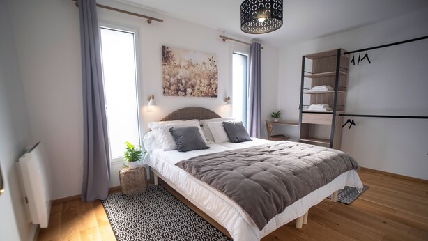 A modern bedroom with a large bed, a bedside table, a wardrobe, and a window with curtains. - Le Jardin des 4 Saisons (Changé)
