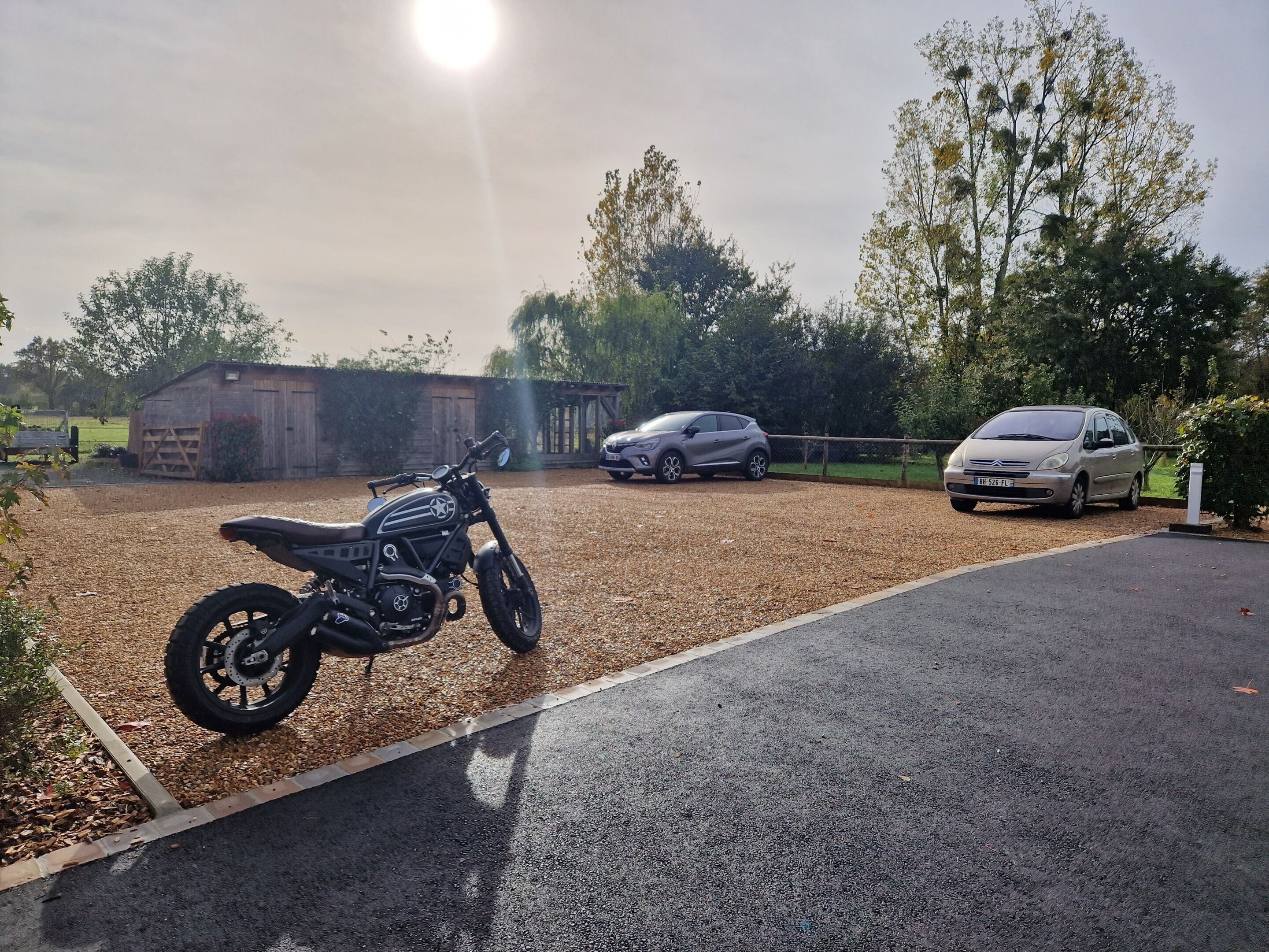 A gravel driveway with a motorcycle parked on the left, a car on the right, and a building in the background.