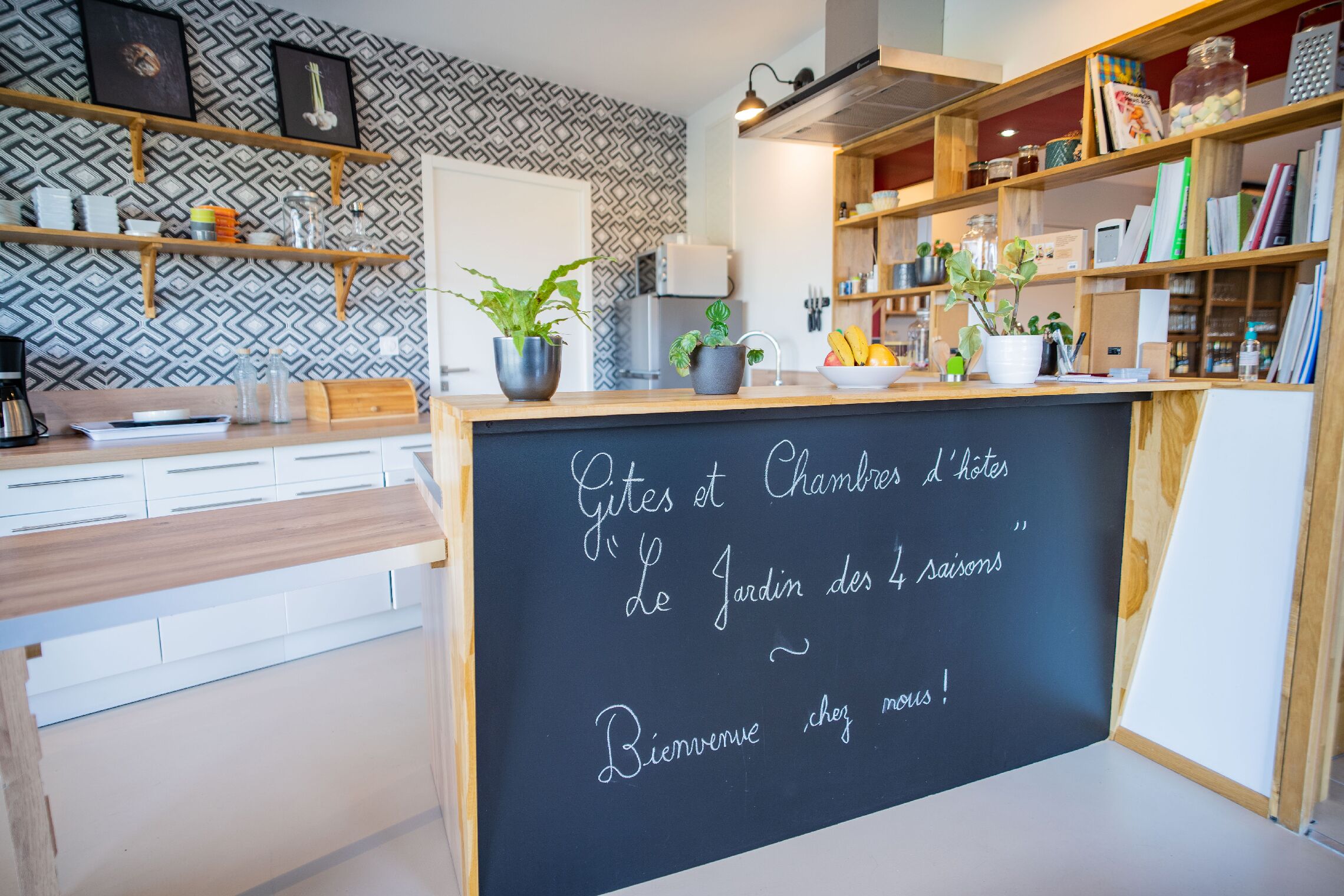 Un espace d’accueil d’hôtel avec un tableau noir affichant un message de bienvenue, des étagères en bois ornées de livres et de plantes, et un mur à motifs.