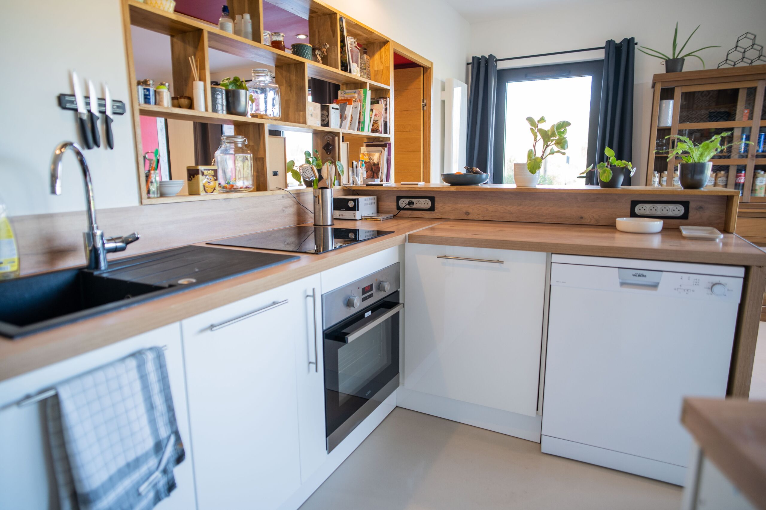A modern kitchen with white cabinets, a wooden countertop, a built-in oven, a dishwasher, and a window with curtains.