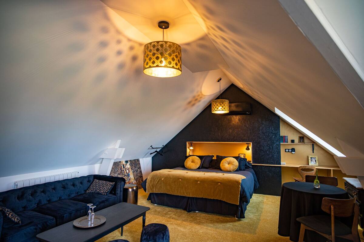 A cozy, modern bedroom with a vaulted ceiling, a large bed with a headboard, a dark blue sofa, and a small table with a lamp.