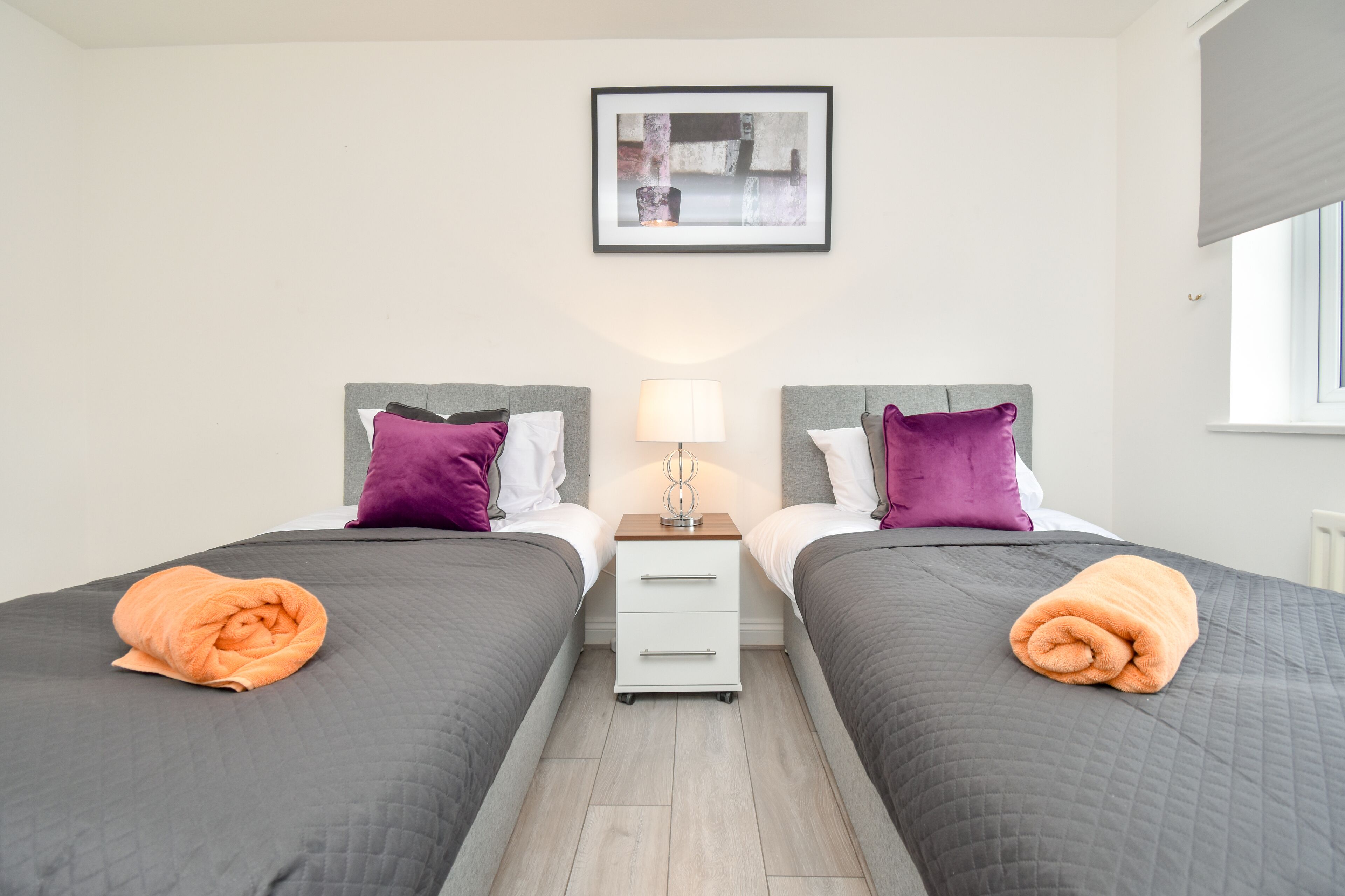A double room with two beds, each with a pillow and a blanket. There is a nightstand with a lamp between the beds.