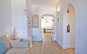 A hallway with a sofa, a small tree decoration, a wooden cabinet, a dining area with a table and chairs, and a fire extinguisher. - The Salty Lodge (Lagos)