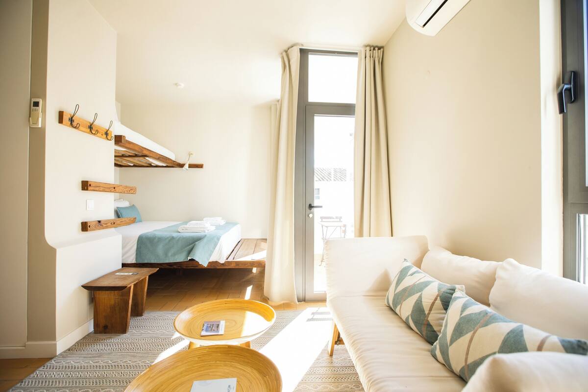 A modern hotel room with a bed, a sofa, a wooden bench, a small table, and a window with curtains.