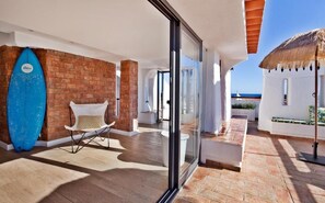 Un espacio exterior moderno con pared de ladrillo, una silla blanca, una tabla de surf azul y un parasol de paja.