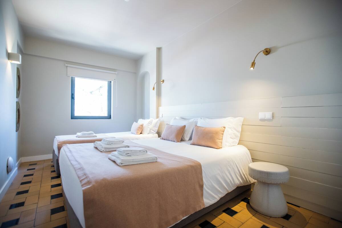 A hotel room with two beds, a window, a bedside table, and a wall-mounted light fixture.