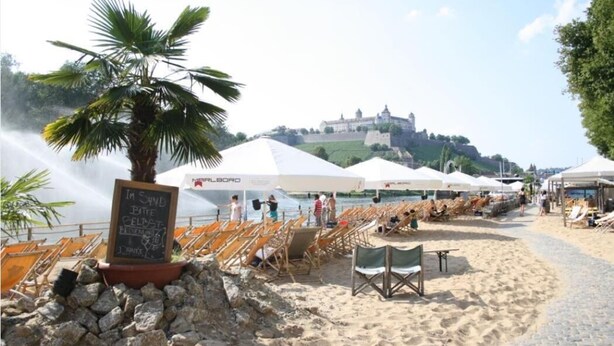 A beach with sun loungers, a palm tree, and a castle in the background. - Wohnen am Ringpark (Würzburg)