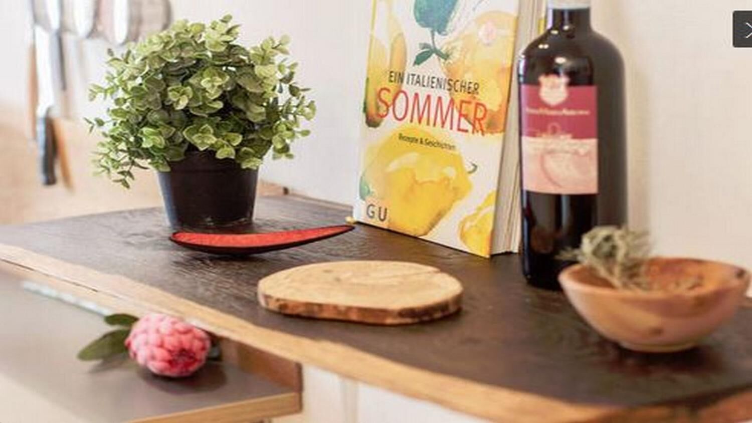 Uma bancada de cozinha com um vaso de planta, um livro intitulado 'EIN ITALIENISCHER SOMMER', uma garrafa de vinho e uma tábua de corte de madeira.