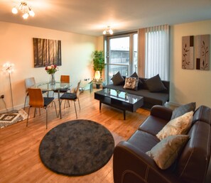 A modern living room with a sofa, coffee table, and chairs. There is a dining table with chairs and a plant. The room has large windows with curtains.