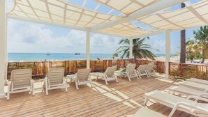 Ein überdachter Außenbereich mit Holzboden und weißen Lounge-Stühlen, mit Blick auf einen Strand mit Palmen und das Meer.