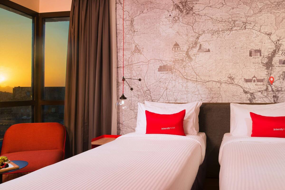 Une chambre d’hôtel avec deux lits, une chaise rouge, une petite table avec des fruits et une grande fenêtre donnant sur un coucher de soleil.