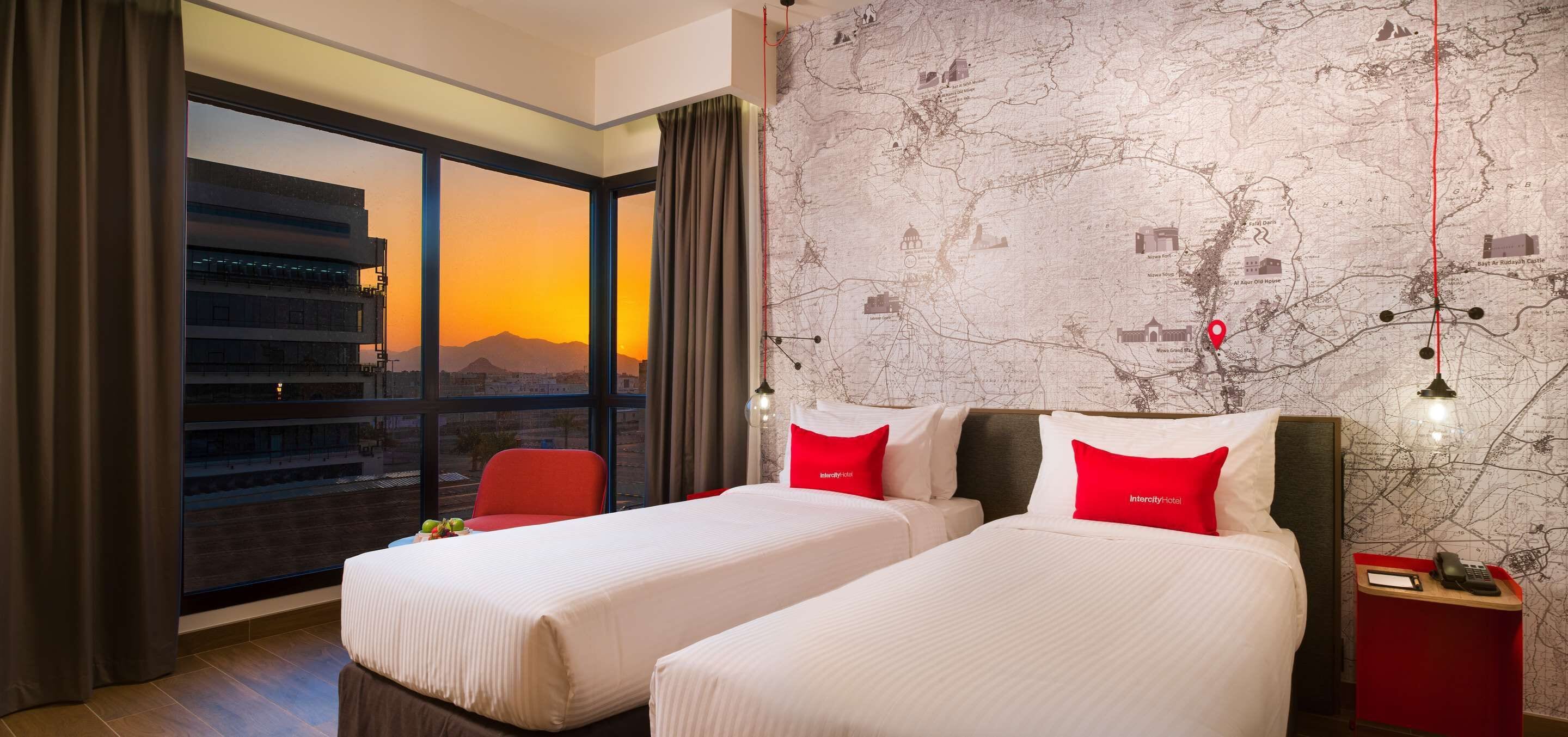 Habitación de hotel con dos camas, un ventanal con vistas a la ciudad, un mapa en la pared y una silla roja.