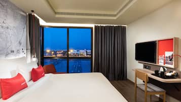 Quarto de hotel com uma cama grande, uma escrivaninha, uma televisão e vista para a paisagem urbana ao entardecer.