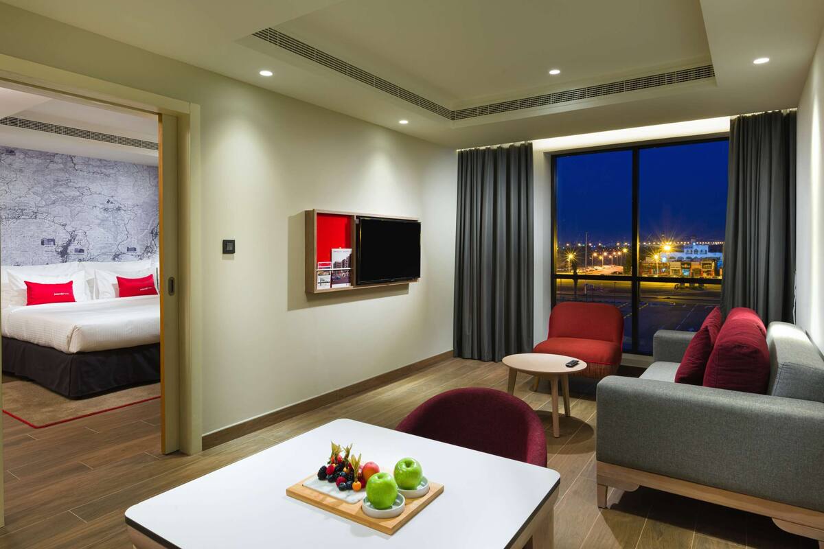 Une chambre d’hôtel avec un lit, une télévision, une table à manger avec des fruits, un canapé et une vue sur la ville la nuit.