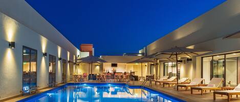 A swimming pool with lounge chairs and umbrellas, surrounded by walls with windows and a bar area.