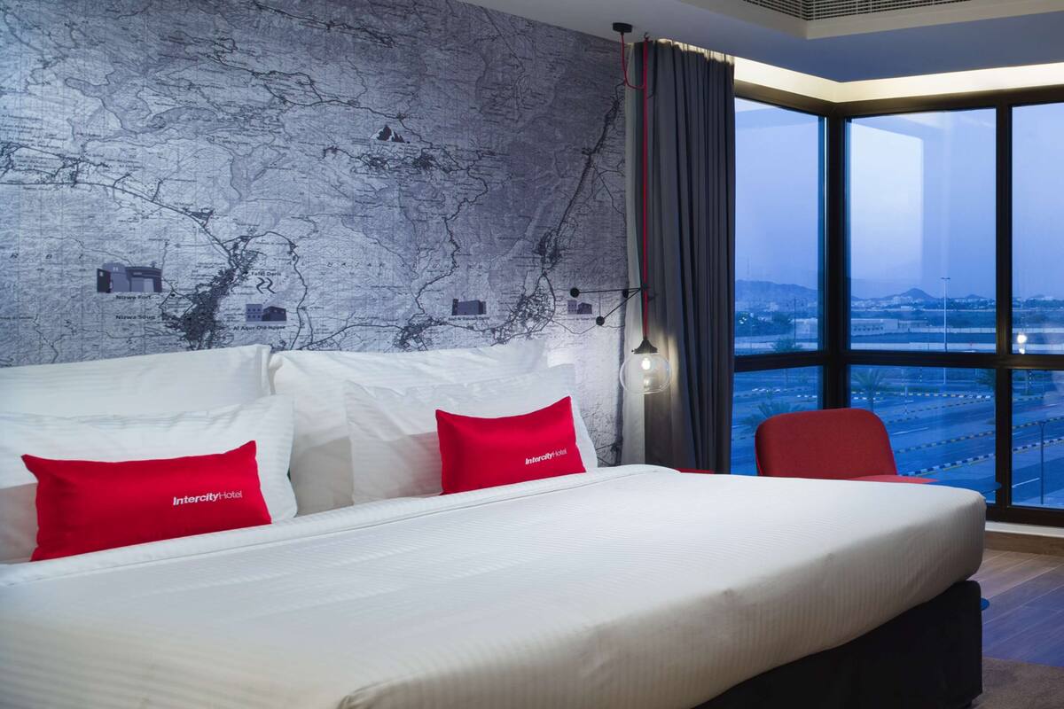 Une chambre d’hôtel moderne avec un grand lit, des oreillers rouges, un mur décoré d’une carte et une vue sur la ville grâce à de grandes fenêtres.
