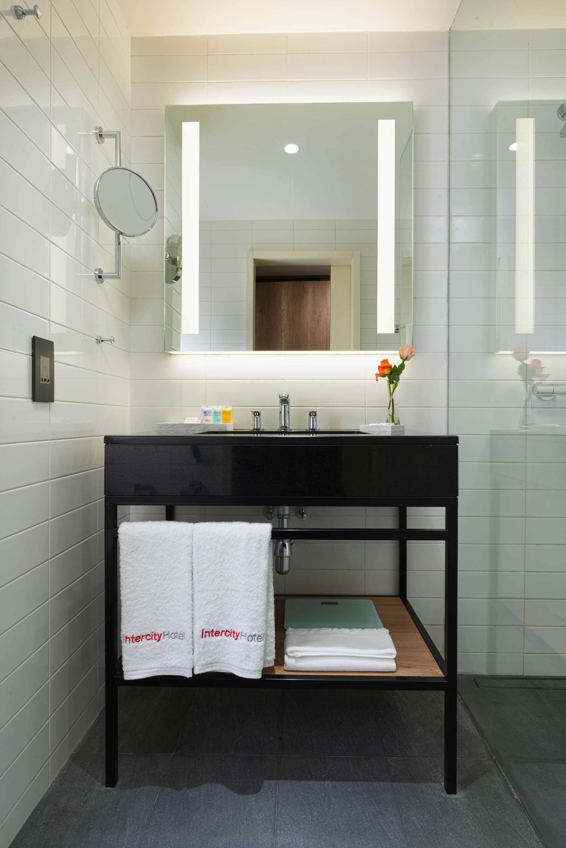 A modern bathroom with a black vanity, white tiles, a mirror, and a small shelf with towels.