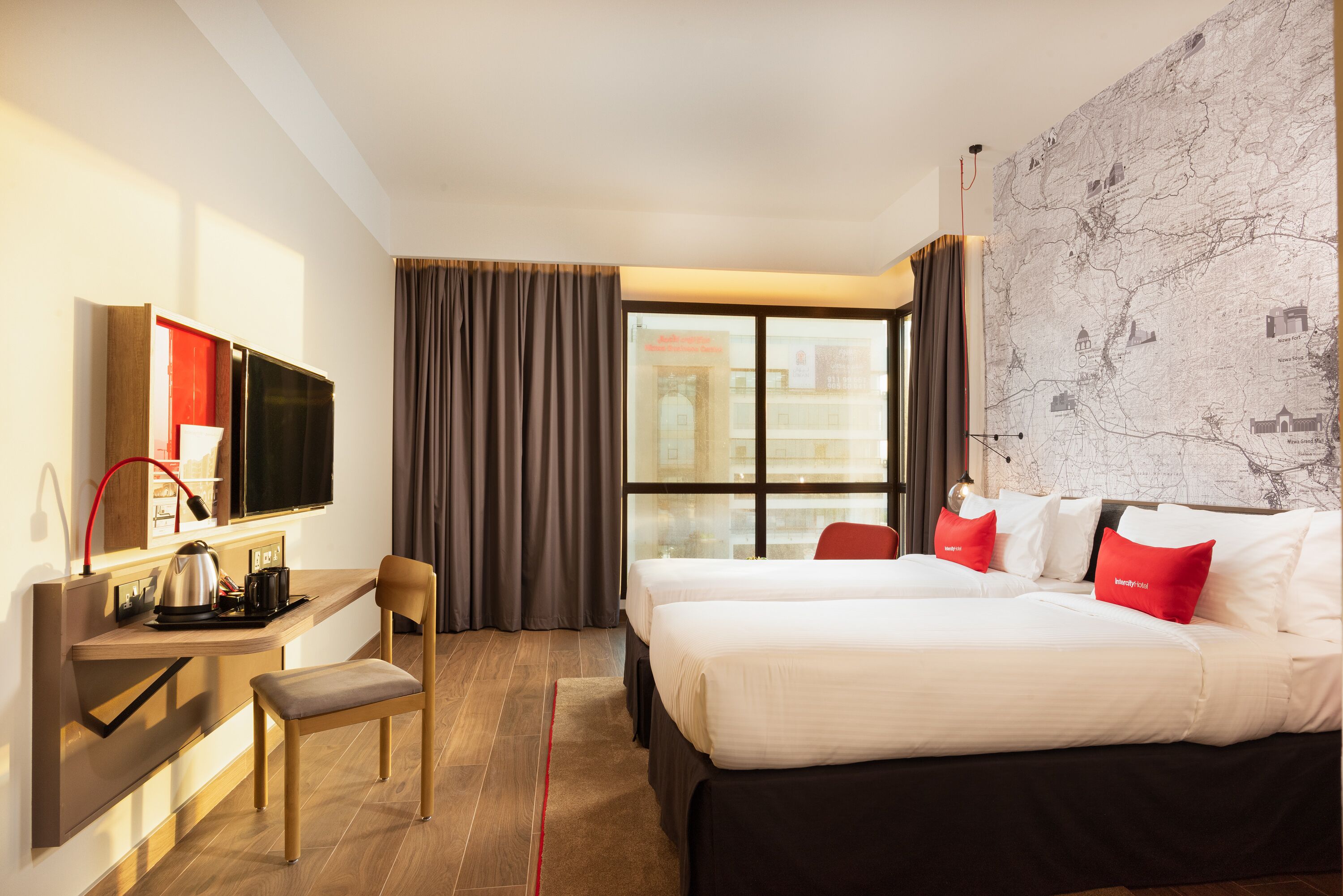 Habitación de hotel con dos camas, un escritorio con televisor, una silla y un amplio ventanal con cortinas.