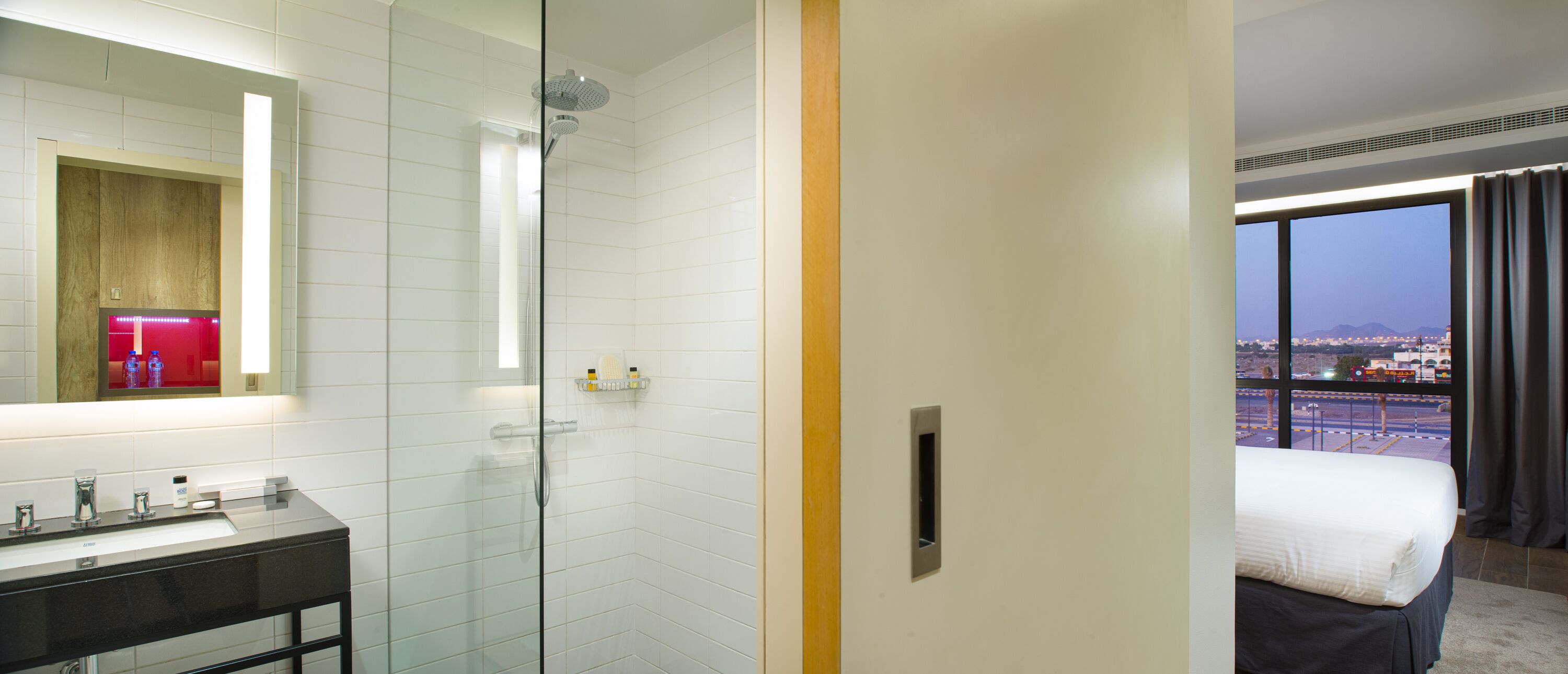 Habitación de hotel con ducha envuelta en vidrio, baño con lavamanos y espejo, y cama con vistas a la ciudad.