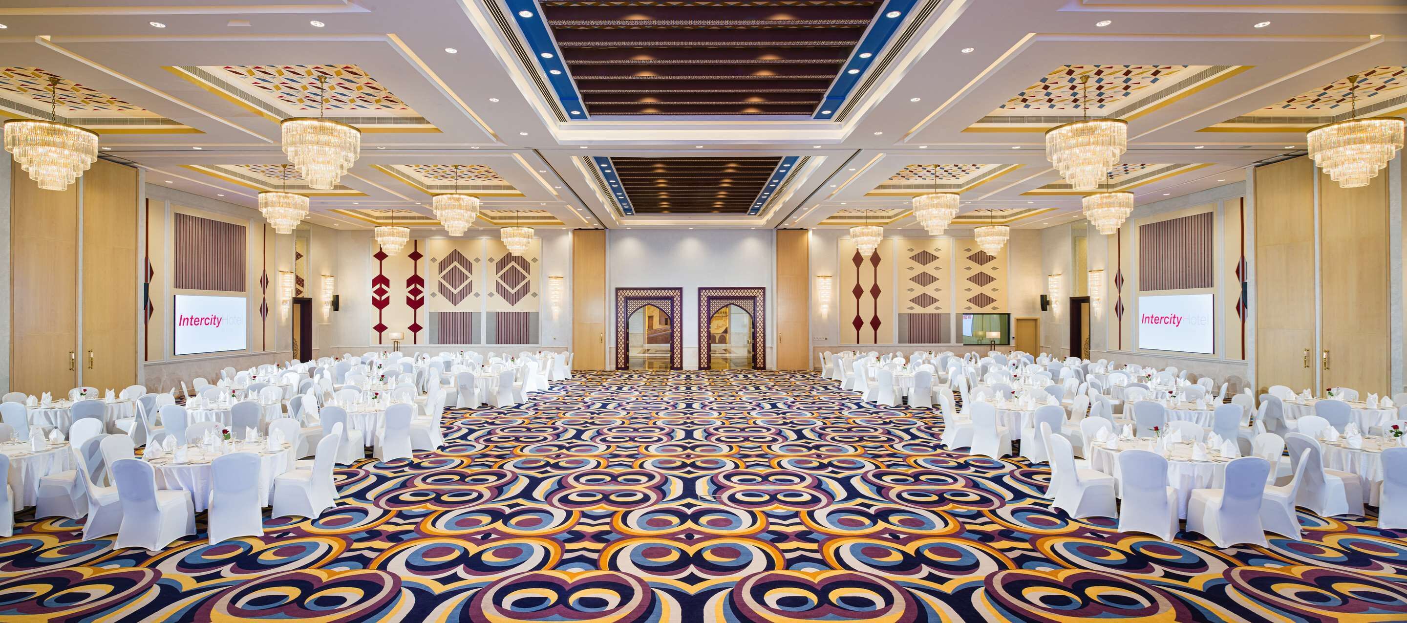 Un salón de banquetes con mesas y sillas blancas, candelabros y alfombra estampada.