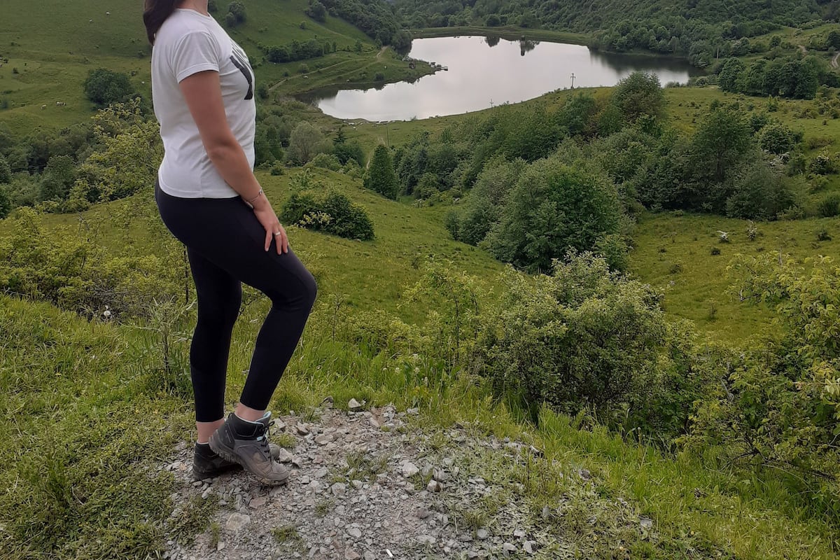 A woman standing on a rocky outcrop overlooking a lake surrounded by lush green hills and distant mountains.