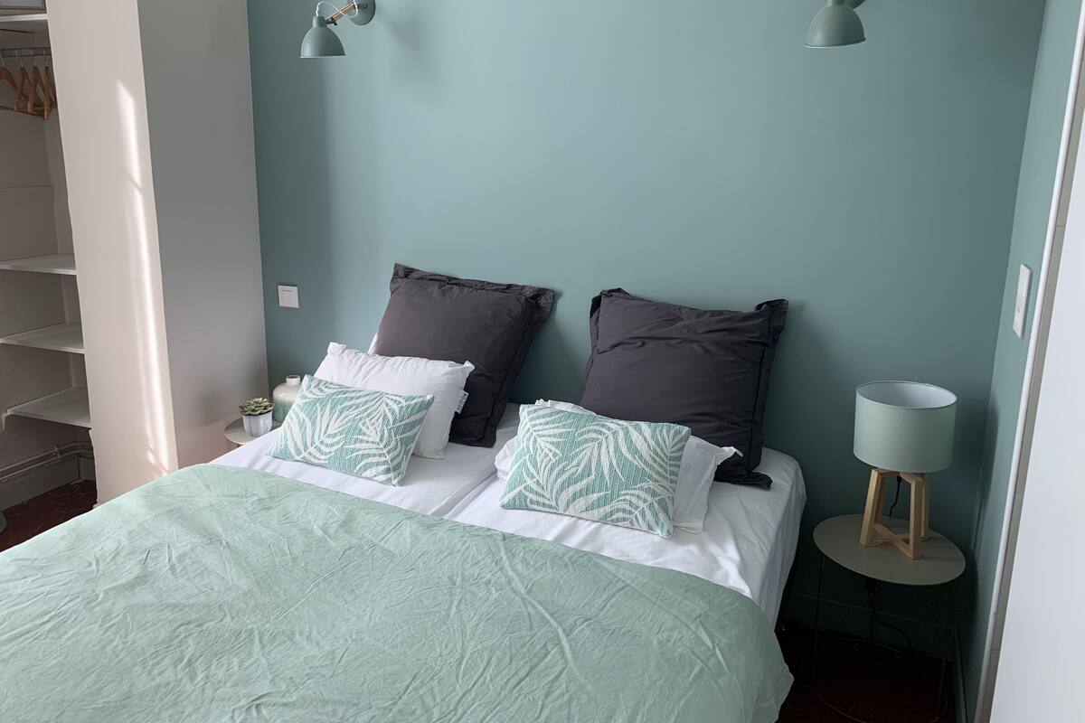 A neatly made bed with a light green bedspread and two pillows, a small bedside table with a lamp, and a wall-mounted shelf with a potted plant.