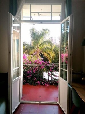 Eine geöffnete Tür führt zu einem Balkon mit Blick auf einen Garten und Palmen.