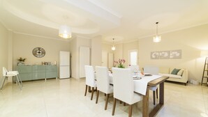A spacious dining area with a wooden table and white chairs, a kitchen with a green cabinet, and a living area with a sofa and a painting on the wall.