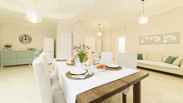 A dining area with a long table set for a meal, a sofa in the background, and a kitchen area to the left.