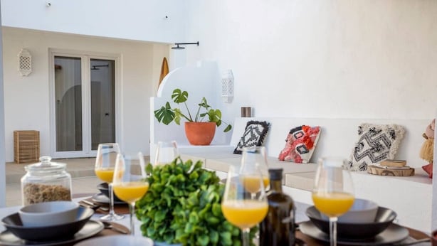Zona pranzo con tavolo apparecchiato per sei, con piatti, ciotole e bicchieri di succo d'arancia. Sono presenti piante in vaso e cuscini decorativi sulla parete.