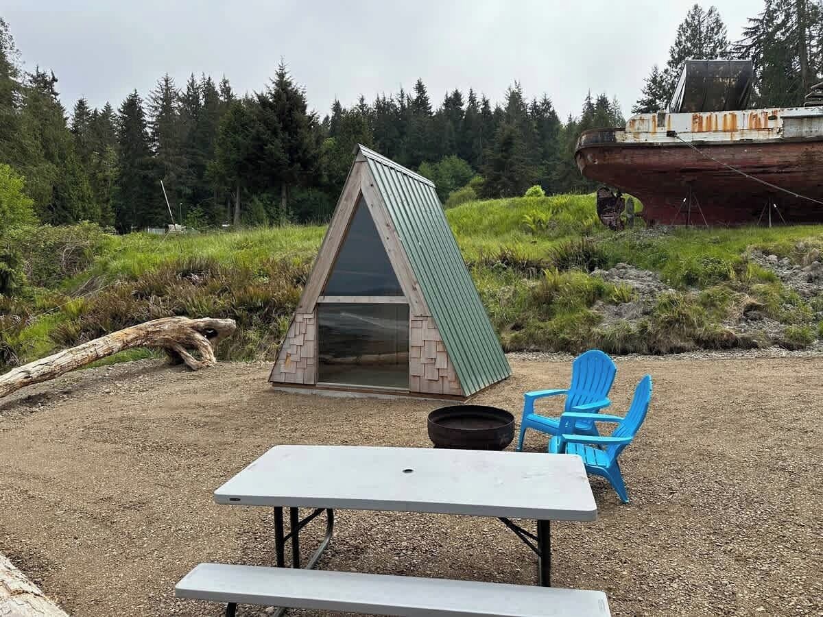 A small, triangular wooden structure with a glass door, a picnic table, and two blue chairs in a natural setting.