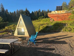A triangular wooden structure with a table and chair in front, surrounded by gravel and greenery.