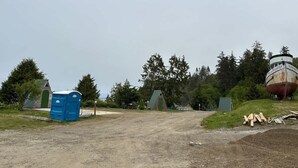 A dirt path leading to a small building with a green door, a blue portable toilet, and a large rusted ship in the background.