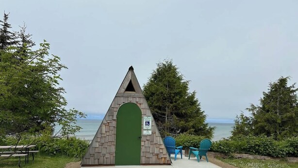 Eine dreieckige Holzkonstruktion mit einer grünen Tür, in Strandnähe mit Stühlen und einer Bank.