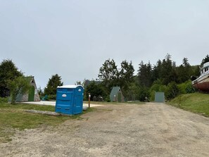 Ein Kiesweg führt zu einer blauen Porta-Toilette, umgeben von Bäumen und kleinen Gebäuden.