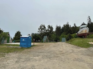 Un chemin de terre mène à un bâtiment doté d’un toit végétalisé et d’un cabinet de toilettes mobiles bleu.