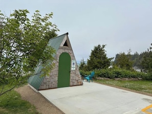 En lille, trekantet bygning med en grøn dør, beliggende nær en belagt plads med en stol og et skilt, der angiver parkering.